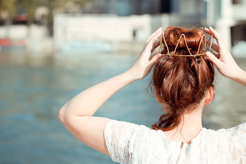 Vue de dos d'une femme aux cheveux roux portant une couronne dorée artisanale, devant un fond aquatique lumineux.
