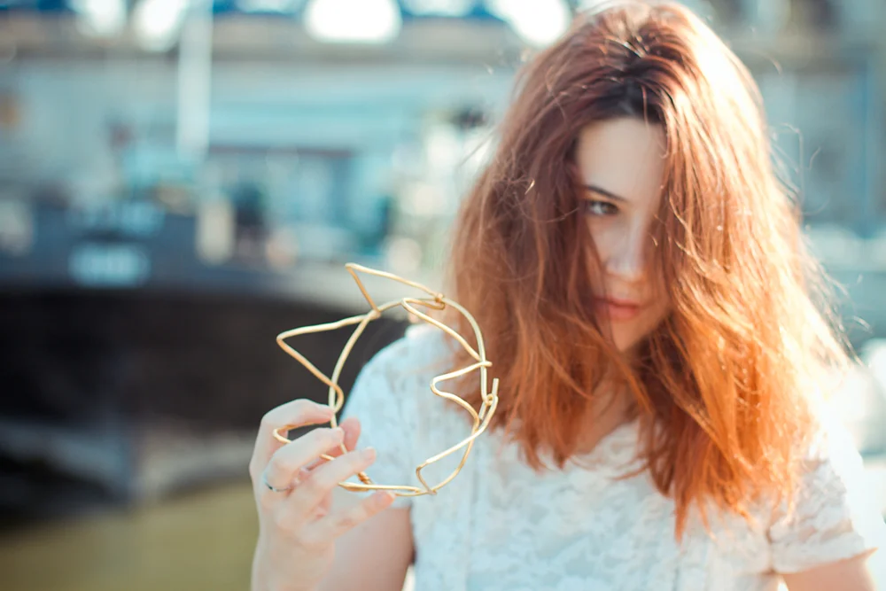 Jeune femme rousse en robe blanche tenant une structure de couronne DIY en métal doré sur un fond flou et lumineux.