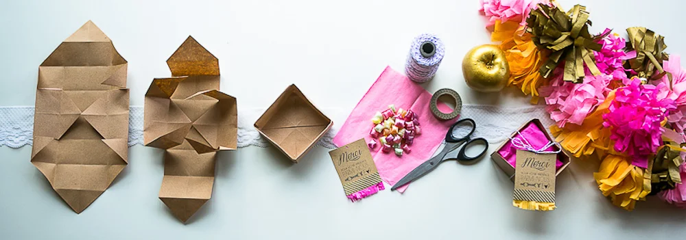 Matériel de papeterie et boîtes en kraft pour DIY, accompagnés de pompons colorés et d'une pomme dorée sur fond blanc.