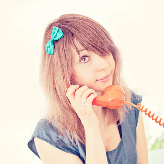 Jeune femme aux cheveux blonds avec un nœud bleu, souriante, tenant un téléphone vintage orange à l'oreille.