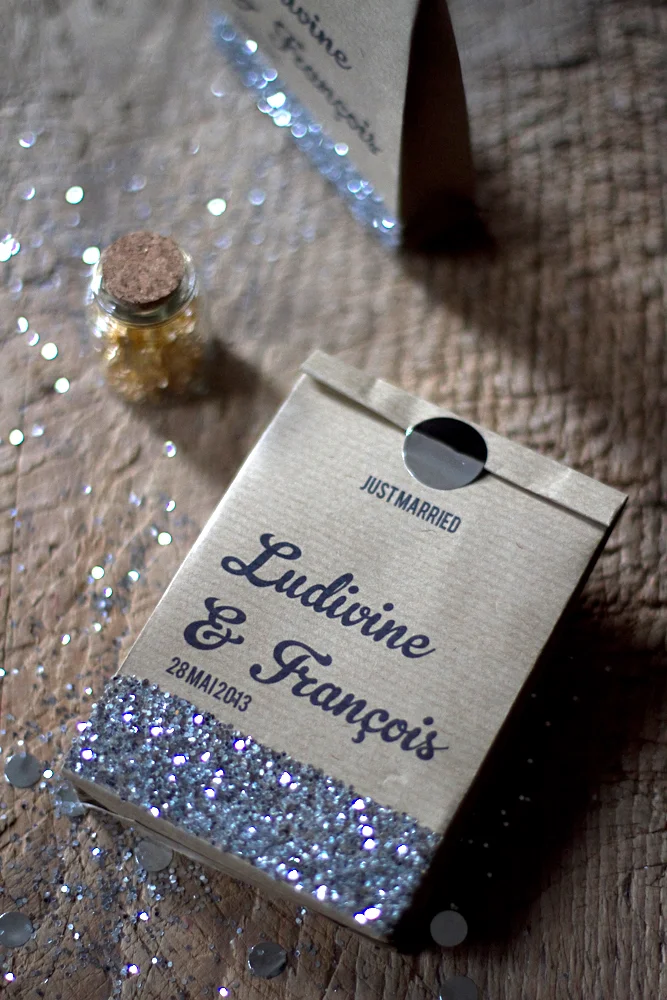 Sachets en papier kraft personnalisés pour mariage, décorés de paillettes argentées sur un fond en bois rustique.