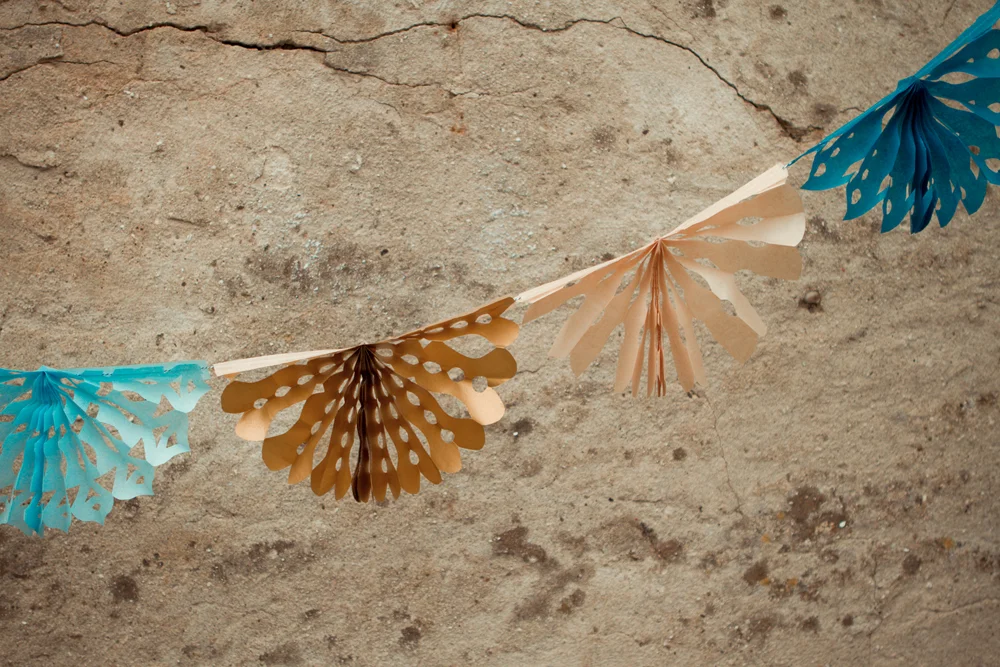 Guirlande de rosaces en papier découpé bleu, beige et doré suspendue devant un mur en pierre texturé.