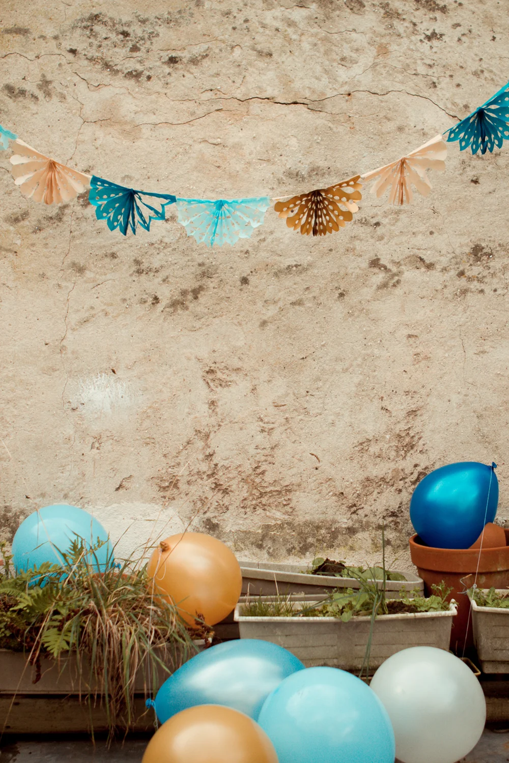 Guirlande de dentelles de papier bleue et beige suspendue devant un mur rustique avec des ballons colorés au sol.