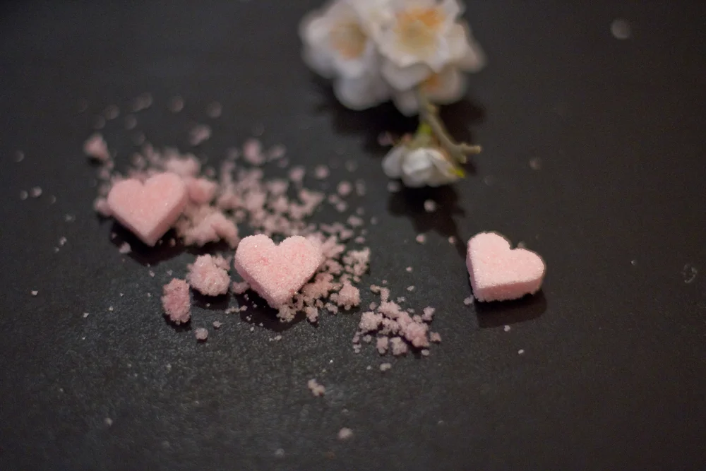 Trois petits sucres roses en forme de cœur et des éclats sur fond sombre avec une fleur blanche floue.