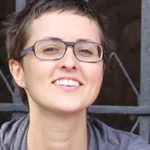 Portrait d'une femme souriante aux cheveux courts et lunettes noires sur un fond sombre.