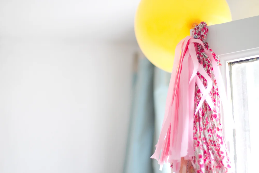 Ballon jaune et gland décoratif rose et fleuri suspendus à un encadrement de fenêtre blanc.