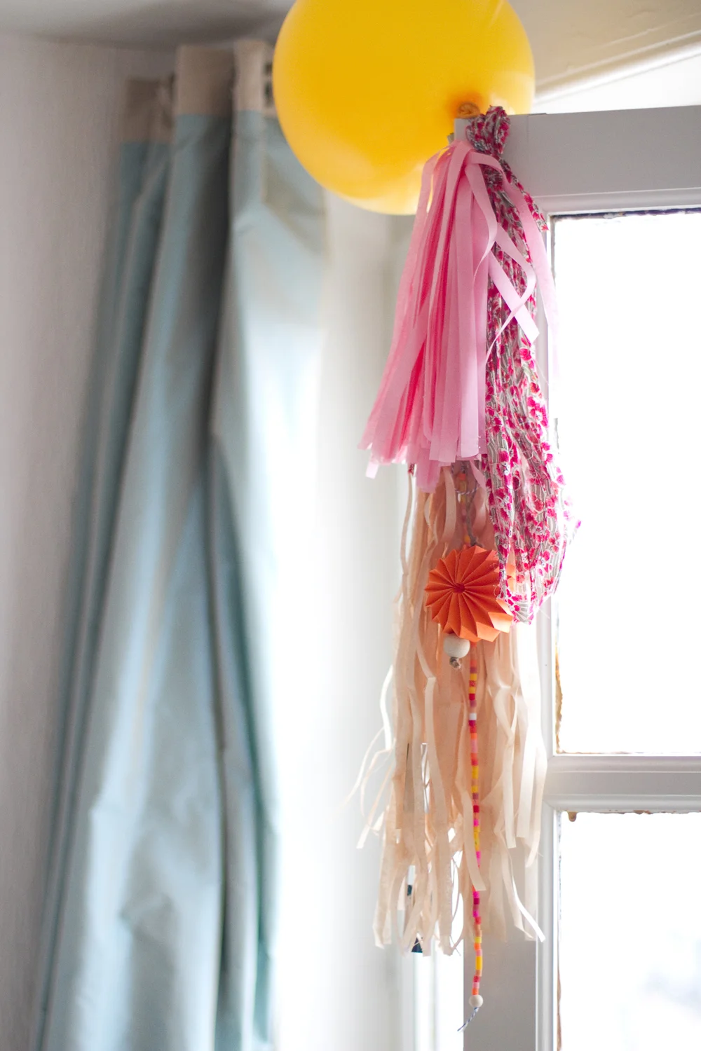 Ballon jaune et glands décoratifs roses et beiges suspendus près d'une fenêtre lumineuse.
