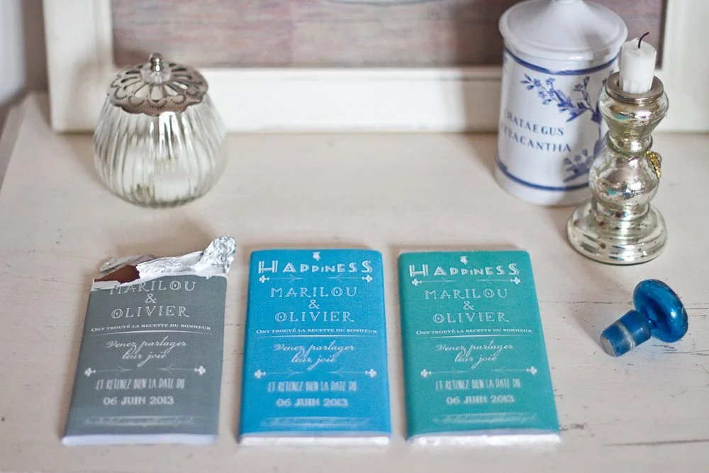 Trois cartons d'invitation bleu canard et gris pour un mariage, posés sur une table blanche avec des objets décoratifs.