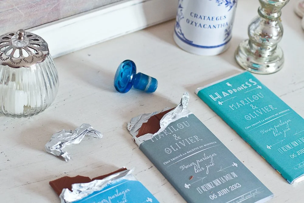 Tablettes de chocolat personnalisées bleues et grises pour un Save the Date, entourées d'objets déco blancs et argentés.