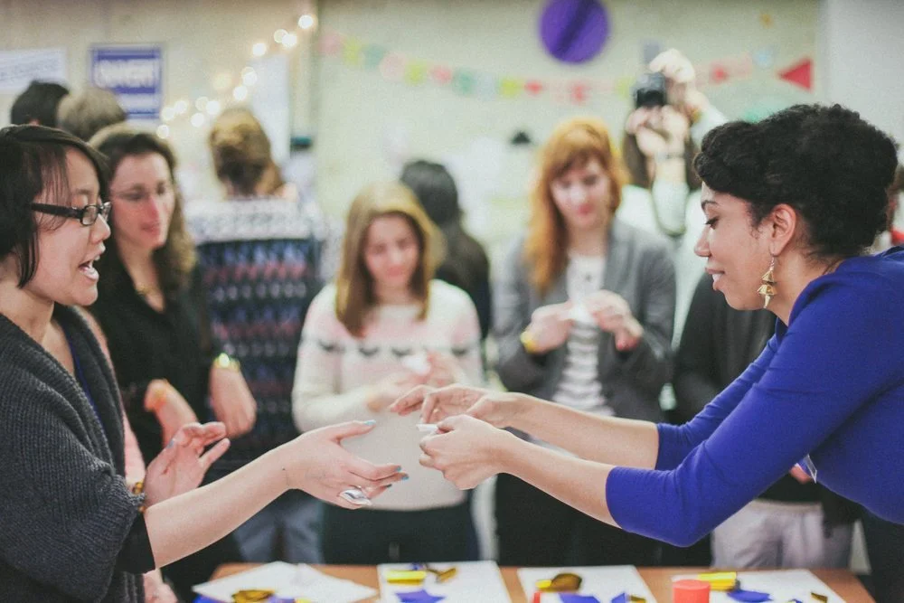 Deux femmes échangent des papiers blancs lors d'un atelier créatif convivial entouré d'autres participants.