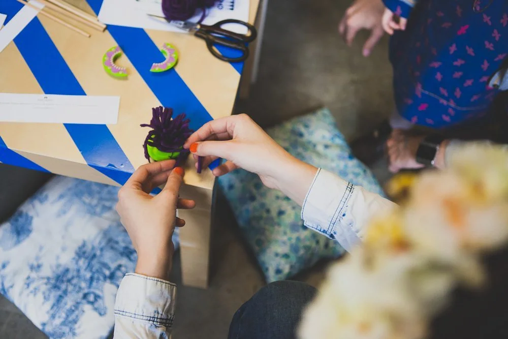 Gros plan sur des mains créant un pompon violet et vert sur une table rayée bleue et blanche.