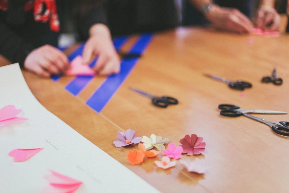 Mains confectionnant des fleurs en papier rose et orange avec des ciseaux sur une table en bois.