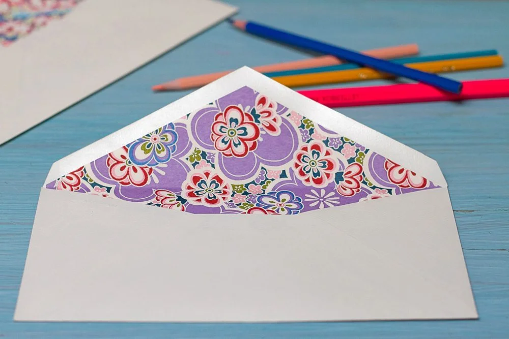 Enveloppe blanche ouverte avec un intérieur floral coloré, entourée de crayons sur un fond bleu.