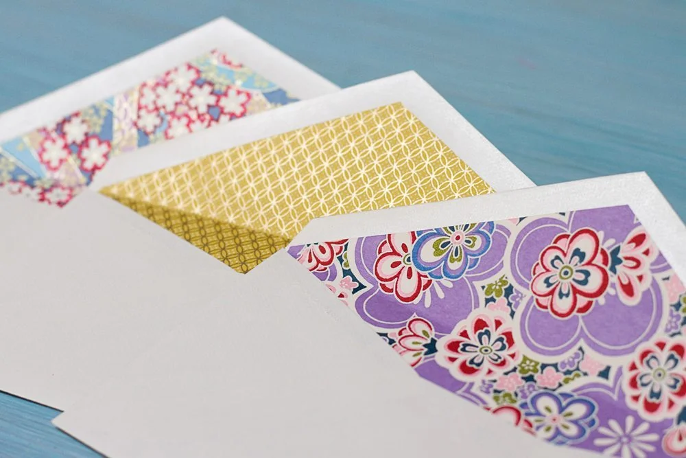 Trois enveloppes blanches doublées de motifs floraux colorés et géométriques dorés sur un fond bleu.