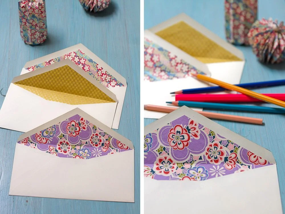 Enveloppes blanches avec doublures colorées à motifs japonais, accompagnées de crayons sur un fond bleu clair.