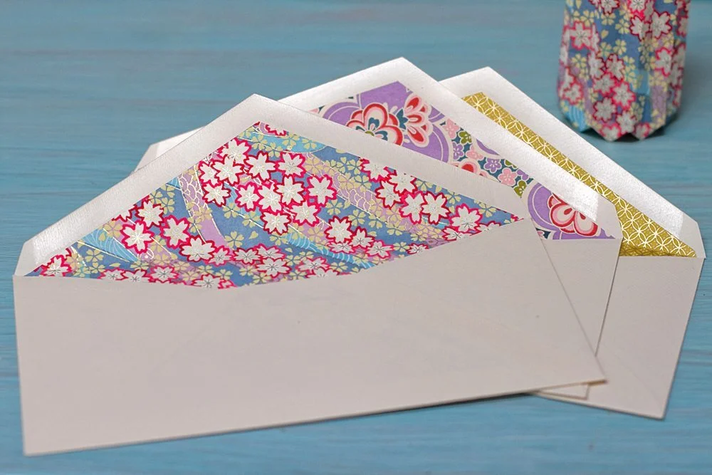Enveloppes blanches avec doublures en papier japonais coloré et motifs floraux sur un fond bleu clair.