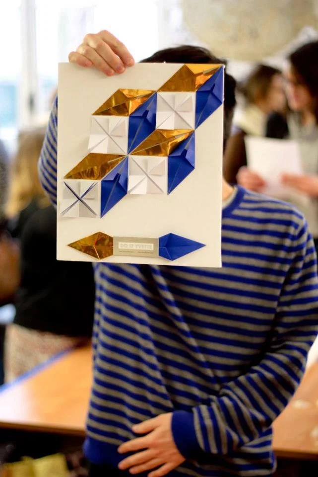 Un homme en pull rayé bleu cache son visage derrière un montage origami géométrique bleu, or et blanc.