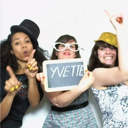 Trois femmes s'amusent avec des accessoires de photobooth, dont une ardoise indiquant YVETTE, sur fond blanc.