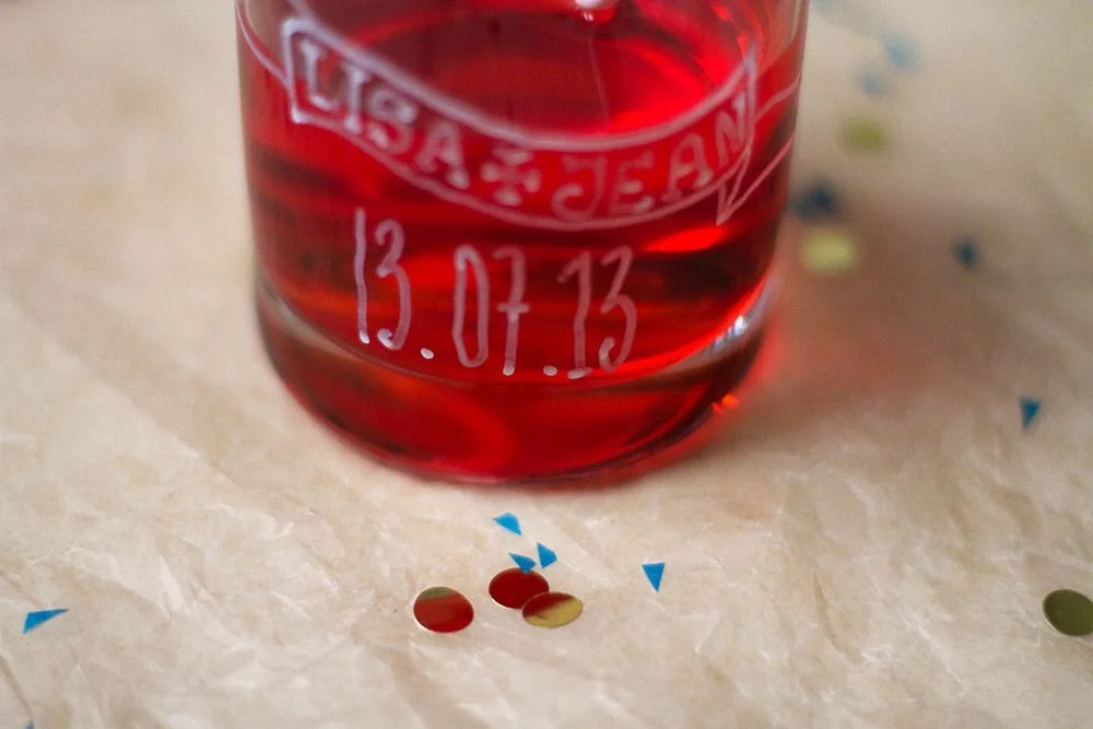 Gros plan d'un verre rouge personnalisé avec des prénoms et une date, entouré de confettis sur un fond clair.