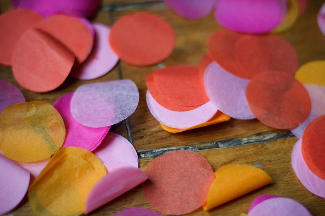Confettis ronds en papier de soie rose, orange et jaune éparpillés sur une table en bois.