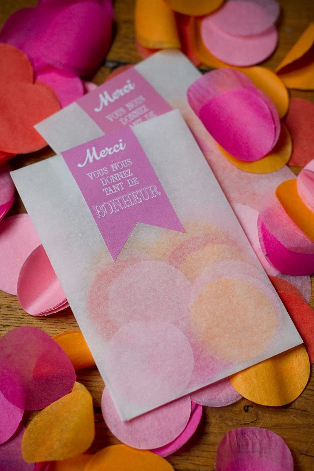 Enveloppes blanches avec rubans roses « Merci » entourées de pétales de fleurs roses et orange sur un fond en bois.