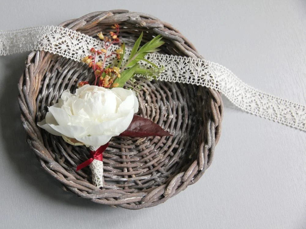Boutonnière blanche et feuillage dans un panier en osier avec un ruban de dentelle blanche sur fond gris clair.