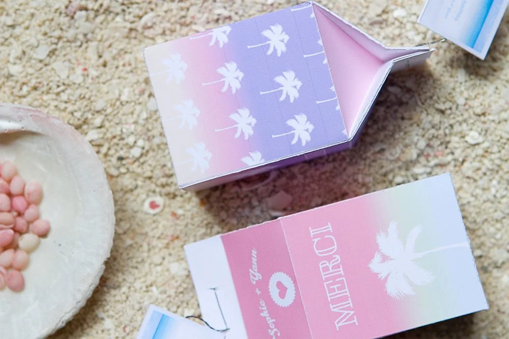 Boîtes de dragées pastel avec motifs de palmiers et inscription Merci sur un fond de sable clair.