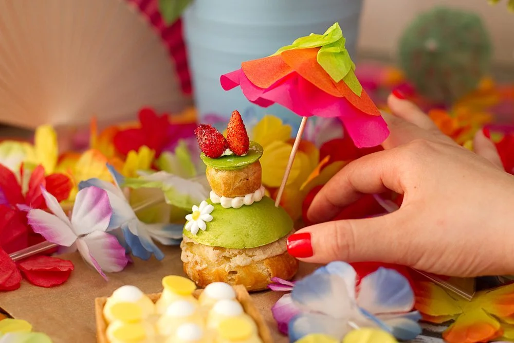 Une main ajuste une petite ombrelle colorée sur un chou à la crème décoré de fleurs et de fraises.