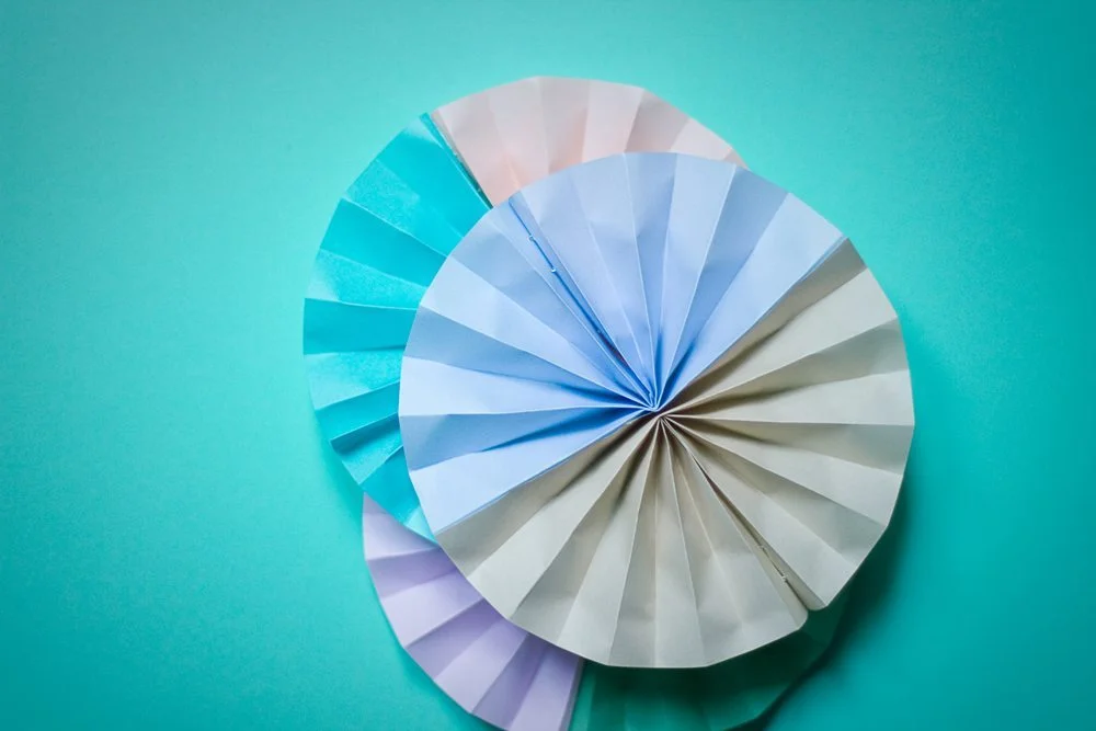 Rosaces en papier plissé bleu, blanc et mauve superposées sur un fond turquoise.