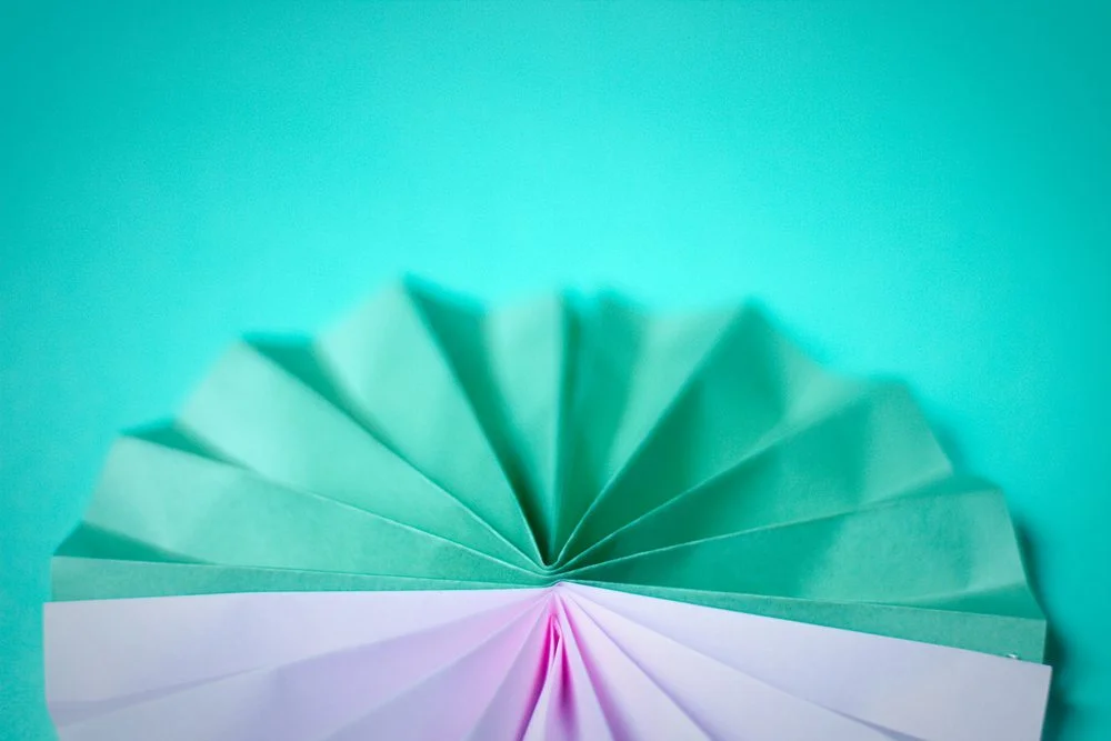 Décoration en papier plié en éventail, dégradé de vert menthe et mauve sur fond turquoise.