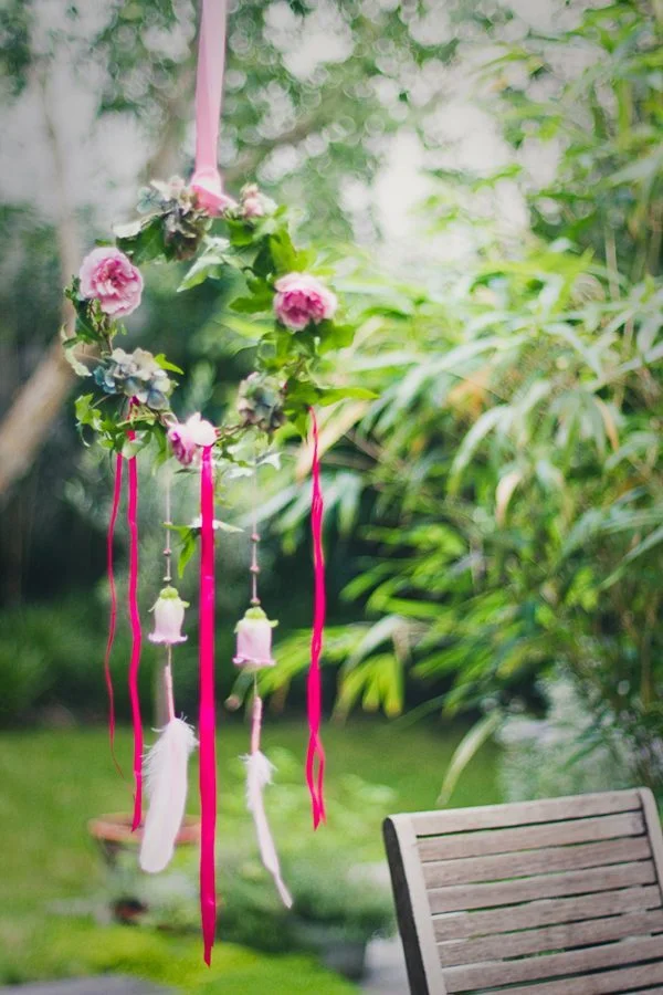 Couronne florale romantique avec rubans roses et plumes blanches suspendue dans un jardin verdoyant.