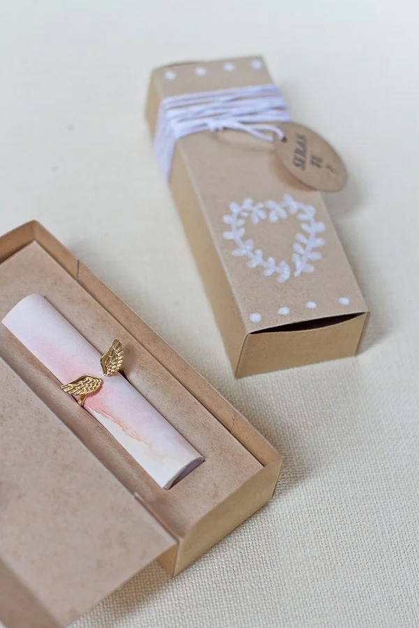 Coffret en carton kraft contenant un rouleau de papier rose pâle orné d'une bague ailée dorée.