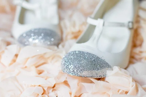 Paire de petits souliers blancs à bout pailleté argenté posés sur un tissu vaporeux couleur crème.