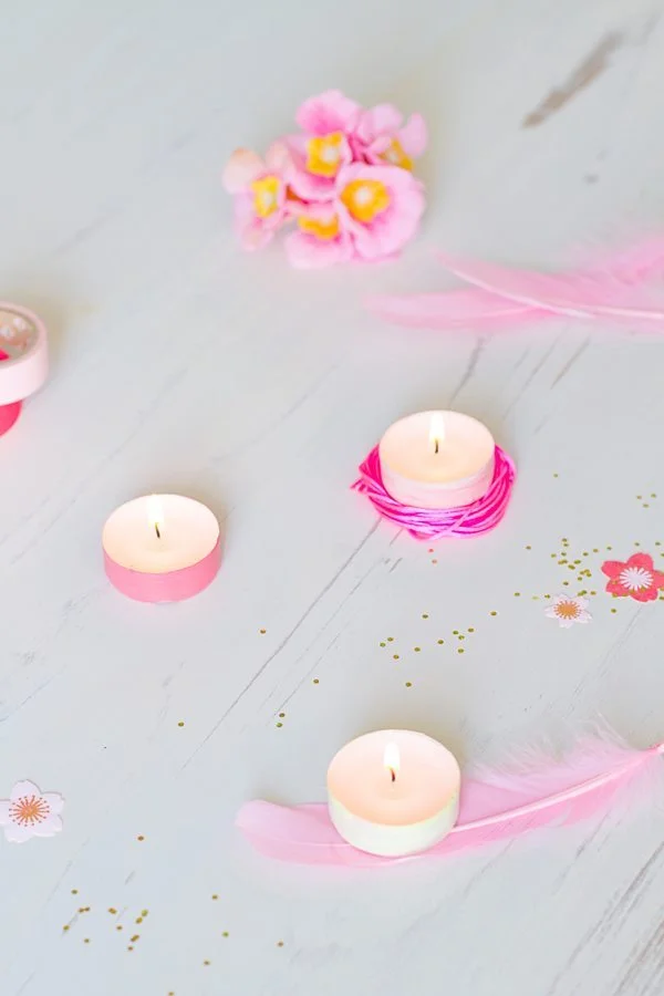 Trois bougies allumées entourées de plumes et de fleurs roses sur un fond blanc lumineux et romantique.