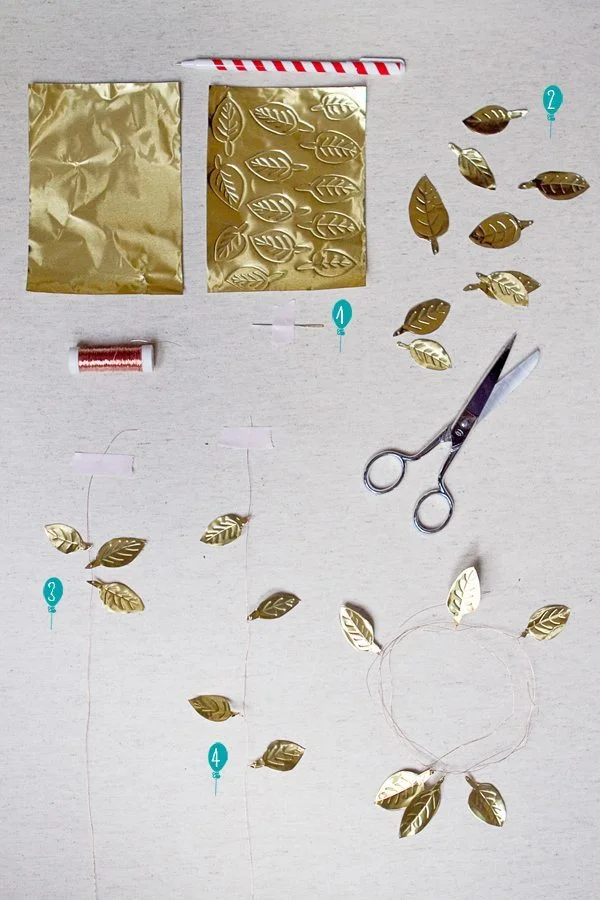 Matériel pour créer une guirlande : feuilles dorées découpées, ciseaux, fil et papier doré sur fond blanc.