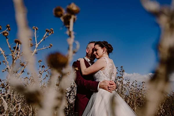 Une mariée en robe blanche et un mari en costume sombre s'embrassent tendrement dans un champ de plantes seche, contre