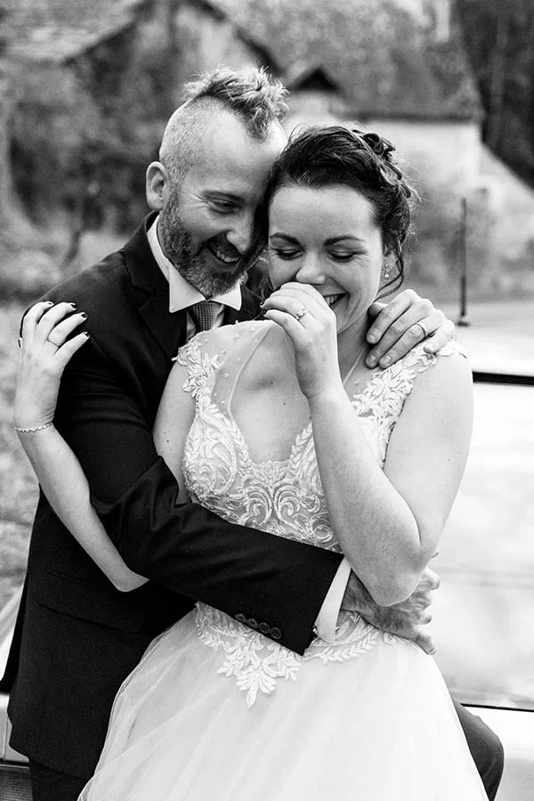 Couple marié complice et rieur en noir et blanc, le marié au style rock et la mariée en robe de dentelle blanche.