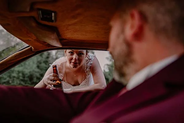 Une mariée en robe blanche et décolleté, tenant une bouteille de bière, regarde un mari en costume de mariage, créant