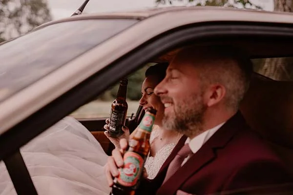 Une mariée et un mari se détendent dans une voiture, partageant des bières avec des bouteilles décorées.