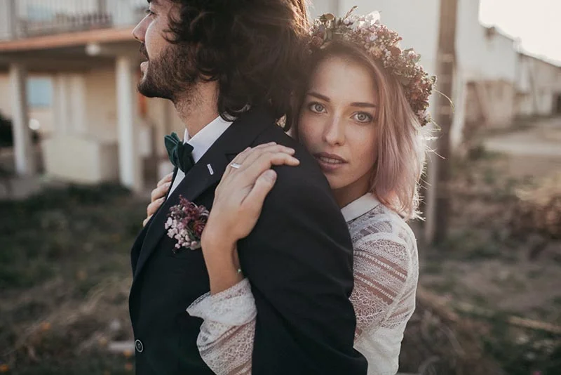 Jeune couple en tenue de mariage folk, la mariée avec une couronne de fleurs embrassant le marié par derrière.