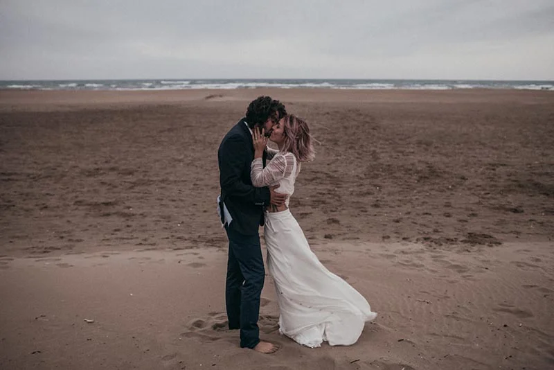 Jeunes mariés s'embrassant sur une plage déserte sous un ciel gris, ambiance folk et romantique.