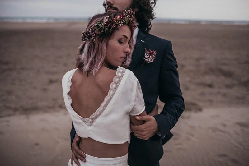 Couple en tenue de mariage folk sur une plage, la mariée portant une couronne de fleurs et un top blanc court.