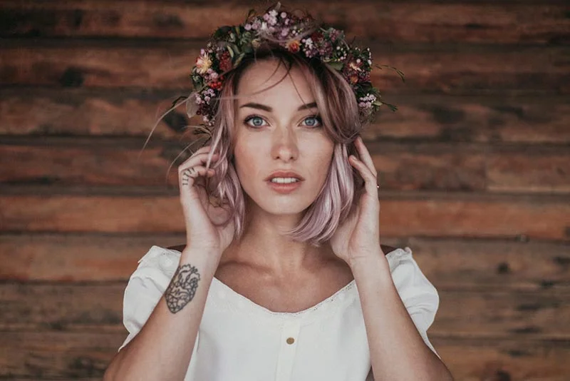 Femme aux cheveux rosés portant une couronne de fleurs sauvages, vêtue de blanc sur fond de bois rustique.
