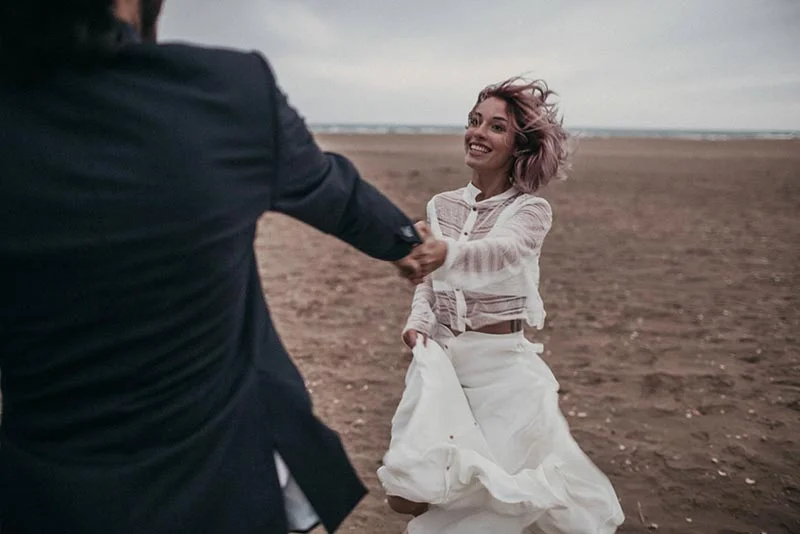 Couple souriant se tenant la main sur une plage sombre, look folk avec robe blanche fluide et costume sombre.