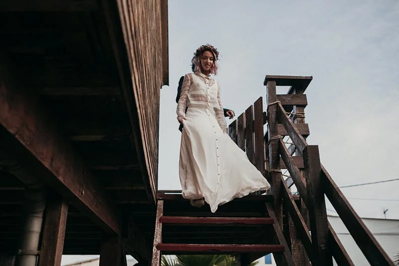 Mariée au style folk en robe blanche et couronne de fleurs, descendant un escalier en bois rustique.