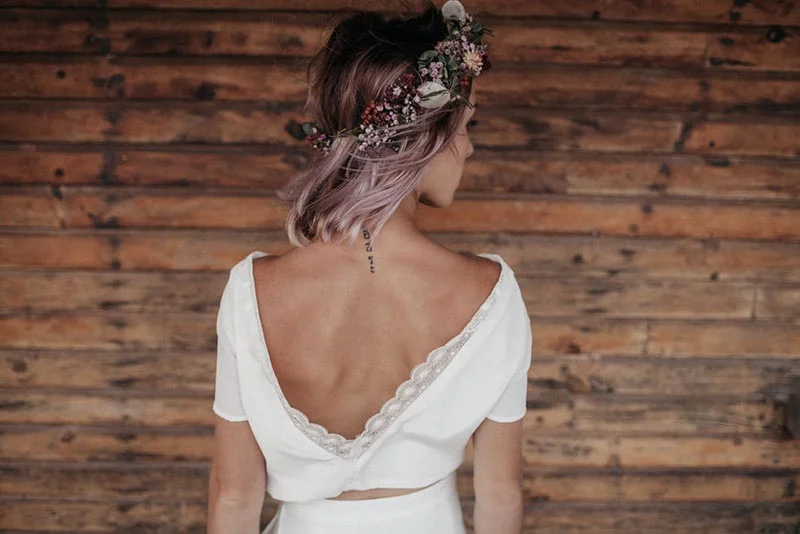 Femme de dos en robe blanche et couronne de fleurs devant un mur en bois, ambiance mariage folk et bohème.
