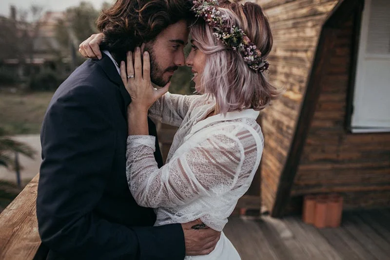Jeune couple marié en tenue folk, s'enlaçant tendrement devant un chalet en bois dans une ambiance romantique.