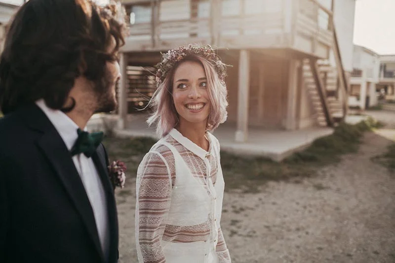 Jeune mariée souriante aux cheveux roses et couronne de fleurs, regardant son époux dans une ambiance folk et lumineuse.