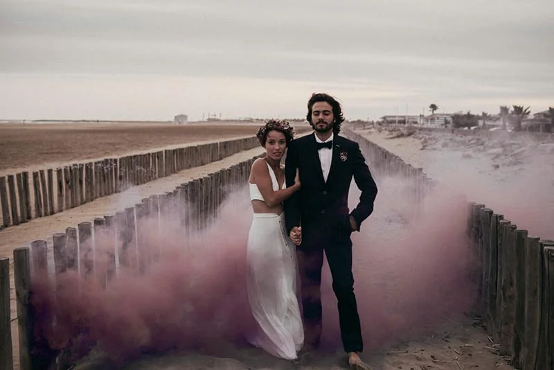 Couple marié élégant posant sur une plage avec un nuage de fumée rose dans une ambiance folk et romantique.