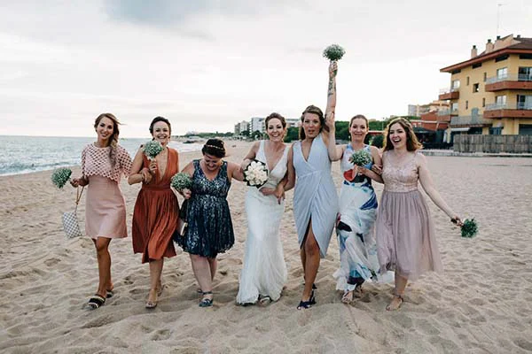Une mariée et ses demoiselles d'honneur sourient et marchent sur une plage de Barcelone avec des bouquets de fleurs.